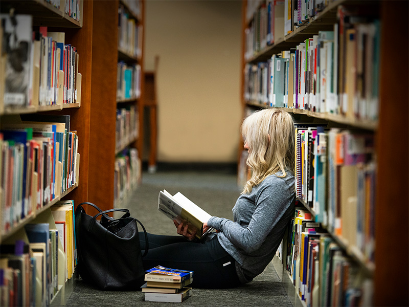 student sitting on floor between library bookcases and reading