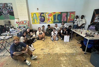 A diverse group of individuals sit and kneel together in an art studio, smiling at the camera. Behind them is a colorful mural spelling out "ASPIRE." Art supplies and completed pieces surround them on tables and walls.