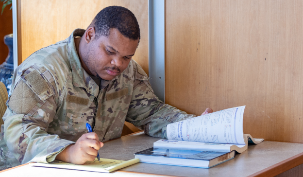 student in military uniform taking notes while reading textbook